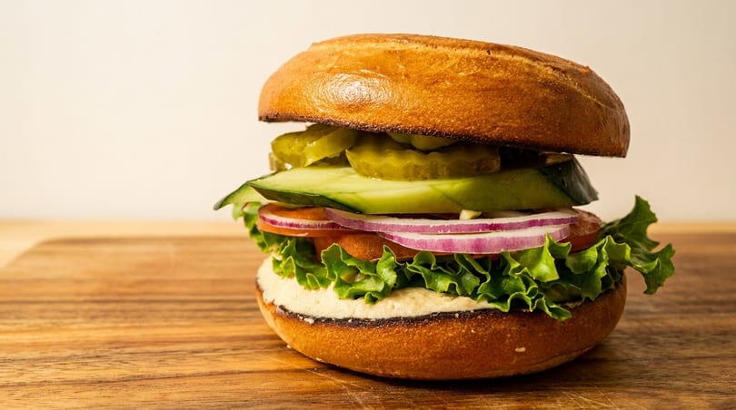 A bagel sandwich with hummus, lettuce, tomato, red onion, cucumber, and pickles on a wooden board.
