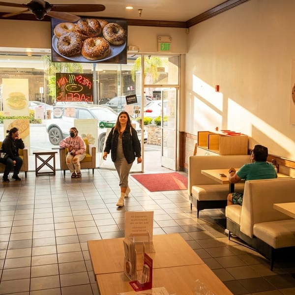 Customers sit and walk through a brightly lit bagel shop with tables, booths, and menu displays.