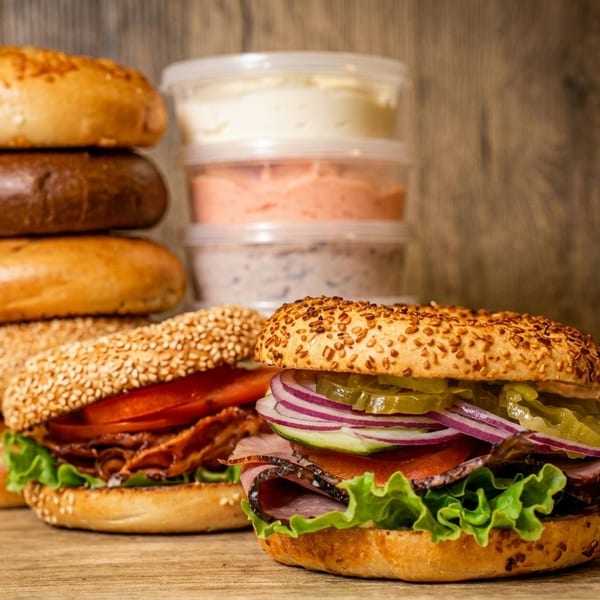 Two loaded bagels, a stack of plain bagels, and three containers of cream cheese on a wooden surface.