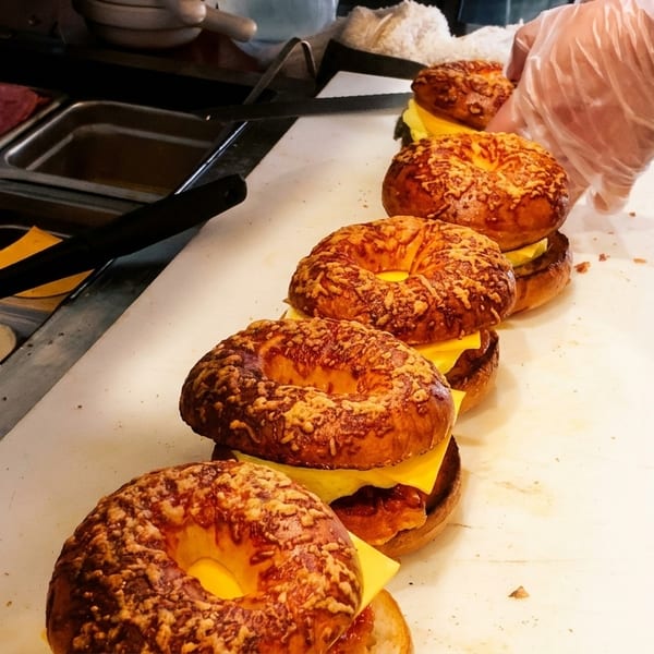 Gloved hand assembling a line of cheesy bagel sandwiches at a deli counter.